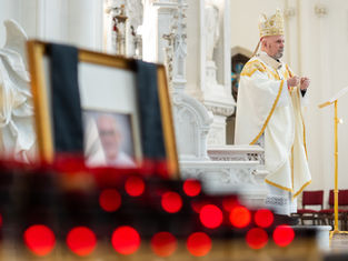 A bishop in ornate white and gold robes stands at a lectern in a church. Red votive candles and a blurred photo are in the foreground.