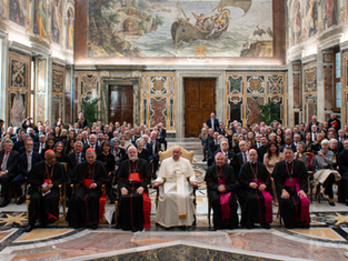 A large group of clergy and attendees sit in an ornate, frescoed room. Central figure in white, surrounded by cardinals in black and red.