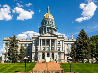 Grand government building with a gold-domed roof, set against a blue sky with few clouds. Surrounded by trees and green lawn.