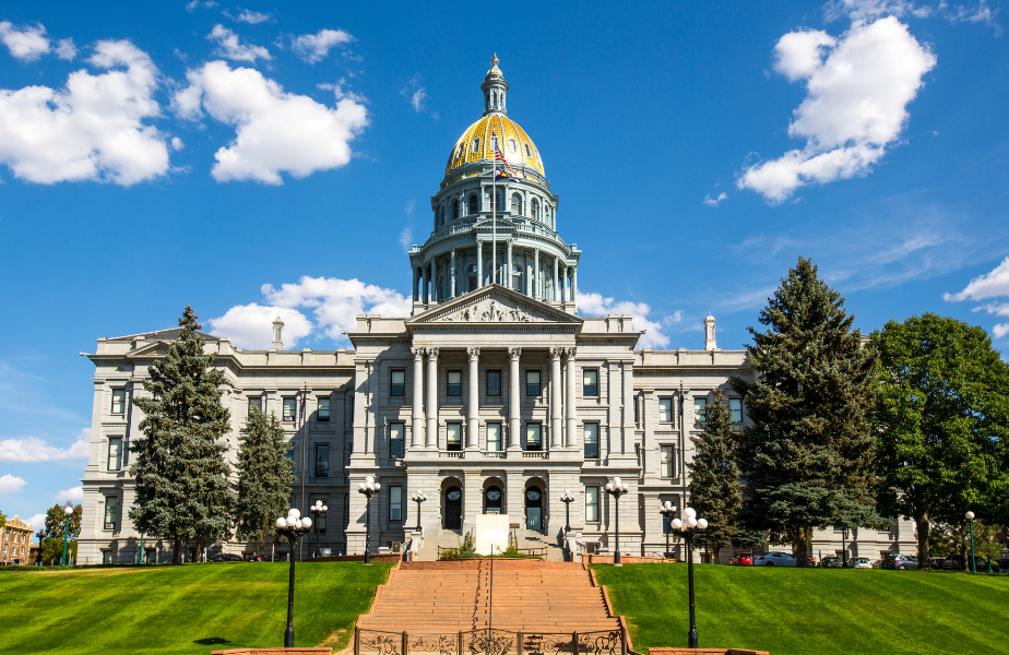 Grand government building with a gold-domed roof, set against a blue sky with few clouds. Surrounded by trees and green lawn.
