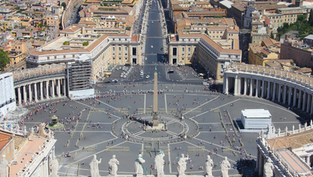 Aerial view of St. Peter's Square, featuring the central obelisk, surrounded by crowds. Colonnades curve on each side, with city buildings beyond.