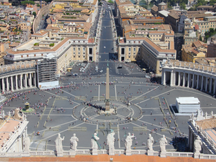 Aerial view of St. Peter's Square, featuring the central obelisk, surrounded by crowds. Colonnades curve on each side, with city buildings beyond.