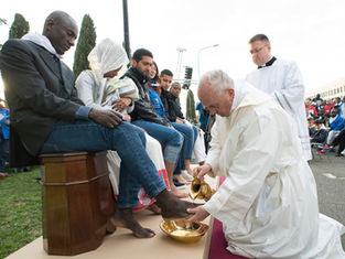 A religious figure in white robes washes a seated person's feet outdoors. A crowd watches. It's a solemn, respectful scene.