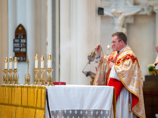 A priest in gold and red robes incenses an ornately decorated altar during a religious service. A marble crucifix and hymn board can be seen in the background. The mood is solemn and prayerful.