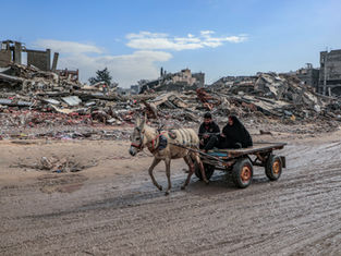 Two people ride a donkey cart through rubble-strewn streets under a blue sky, conveying a sense of resilience amid destruction.
