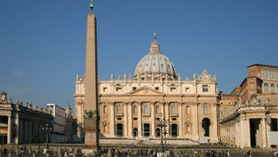 St. Peter's Basilica with a tall obelisk in front under a clear blue sky. Tourists gather in the square, capturing a historic and serene scene.