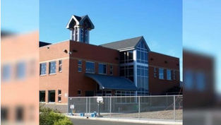Red-brick building with large glass windows and tower against blue sky. Fenced yard in front, suggesting a secured area or campus.