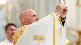 A priest in white and gold vestments holds up a communion wafer during a religious ceremony inside a church, focused and reverent mood.