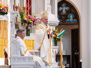 A bishop in ornate robes speaks at a church altar with flowers; another priest is seated nearby. Stained glass and yellow drapes are seen.