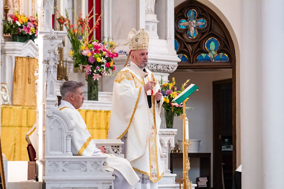 A bishop in ornate robes speaks at a church altar with flowers; another priest is seated nearby. Stained glass and yellow drapes are seen.