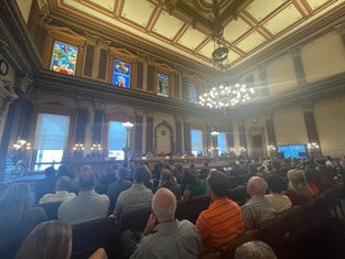 Large audience in a grand hall with stained glass windows and chandeliers, facing a panel. Mood is formal and attentive.