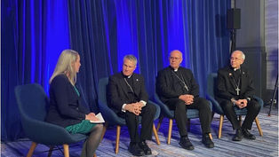 A woman interviews three men in clerical attire seated on blue chairs, against a blue curtain backdrop. The setting is formal and calm.