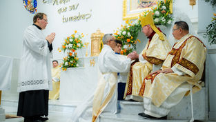 Catholic clergy in white and gold vestments share a blessing at the altar; Spanish wall text reads No estoy yo aquí que soy tu madre?