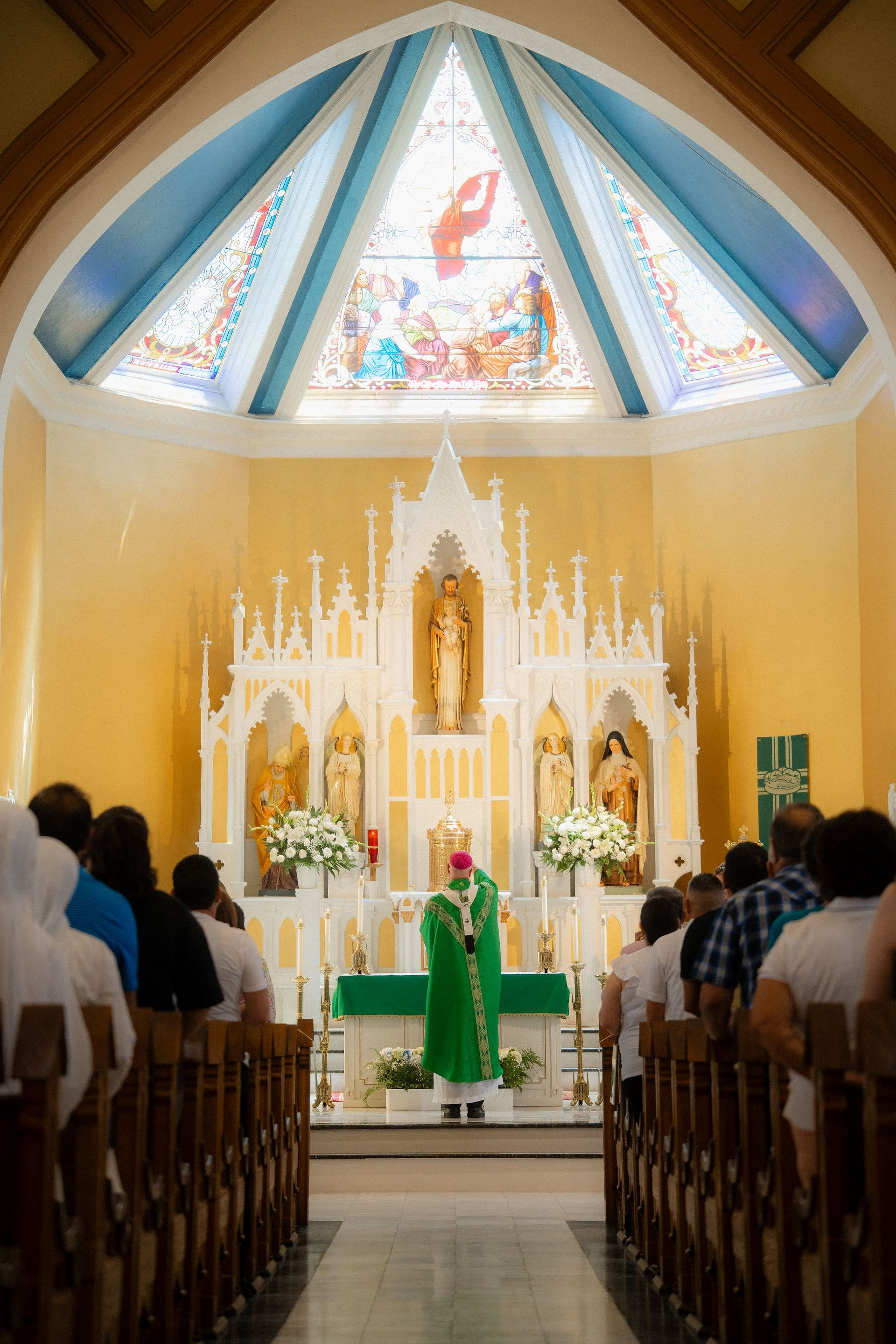 Catholic mass in a bright church: a priest in green faces the altar as congregants stand beneath stained glass and a white altar screen.