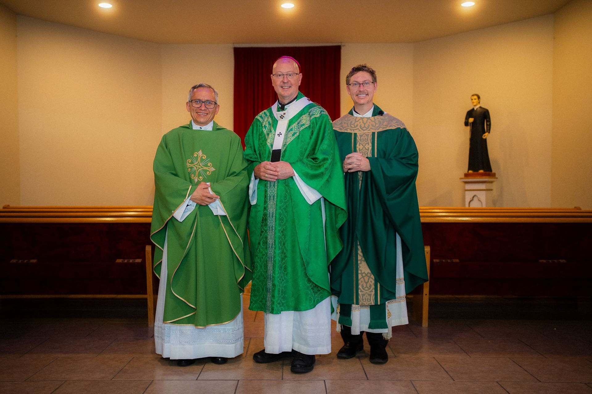 Two smiling priests and one smiling bishop in green vestments stand in a chapel before a red curtain, with a saint statue in the background.