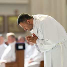 A man in a white robe bows in prayer during a church service, with a blurred seated congregation in the background.
