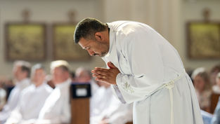 A man in a white robe bows in prayer during a church service, with a blurred seated congregation in the background.