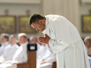 A man in a white robe bows in prayer during a church service, with a blurred seated congregation in the background.