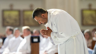 A man in a white robe bows in prayer during a church service, with a blurred seated congregation in the background.