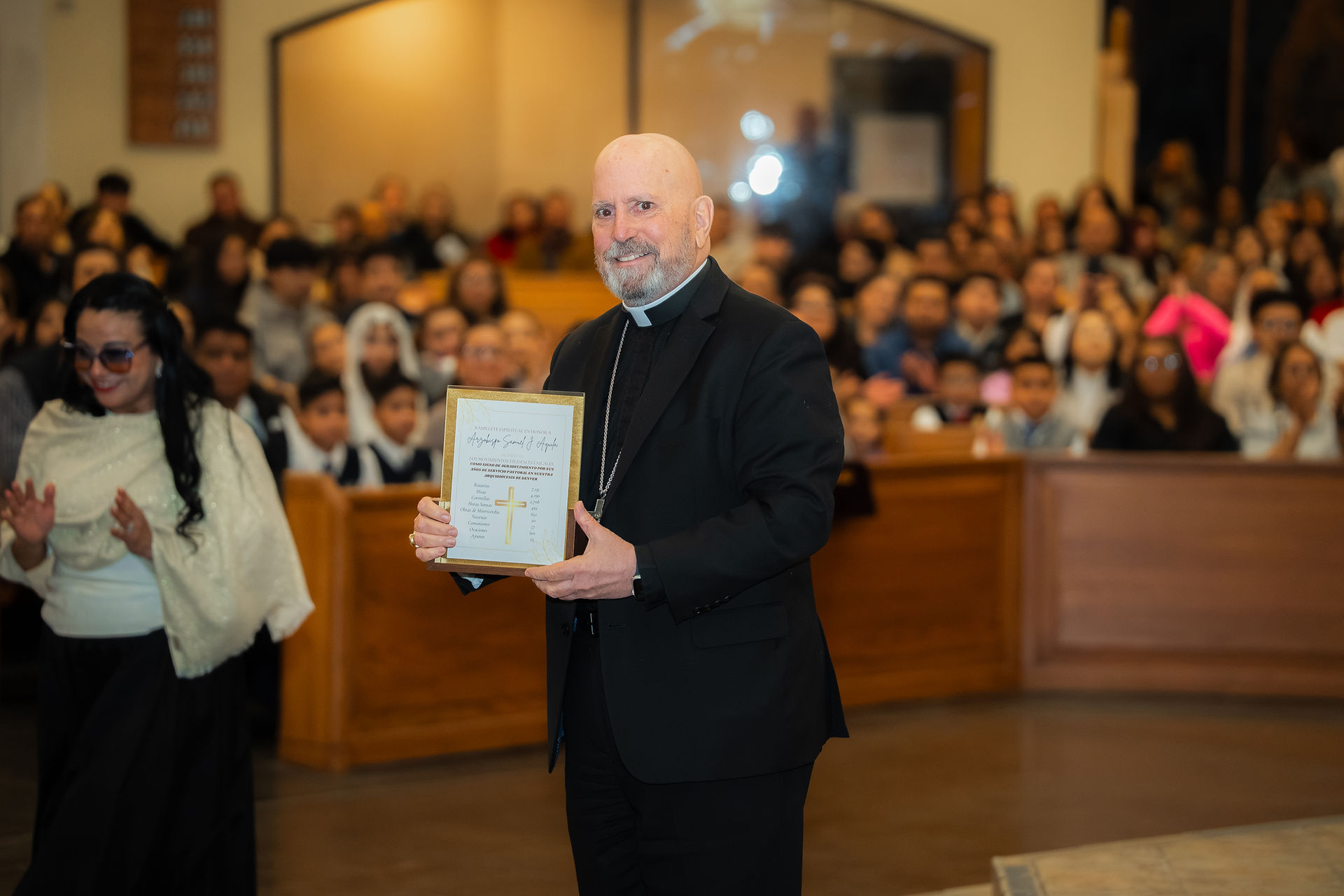 Man in black suit holds a framed certificate with a gold cross in a crowded church. Joyful atmosphere, woman in white nearby.