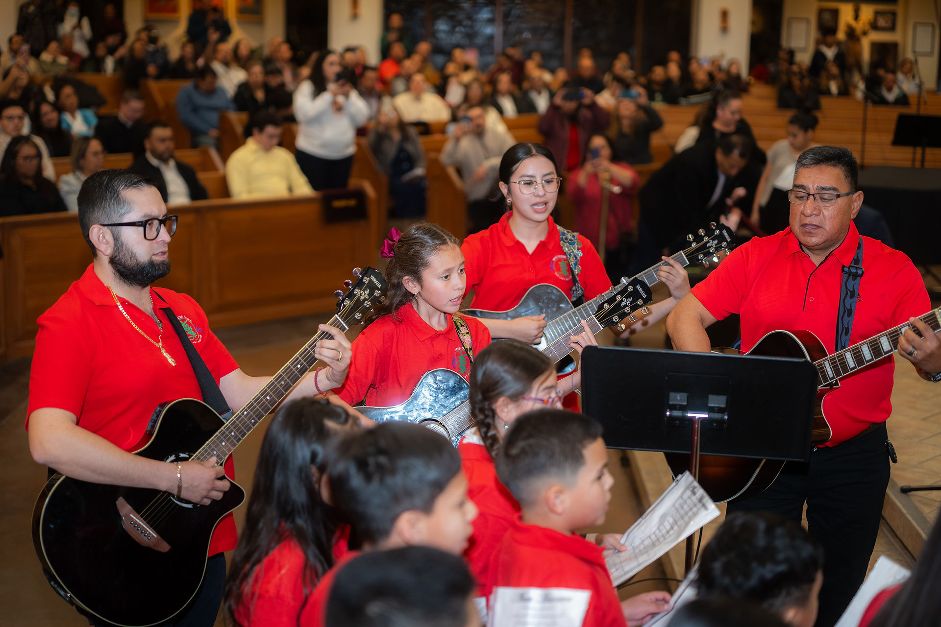 Several musicians, some young, some older, sing and play instruments, wearing red shirts. A congregation is visible in the background.