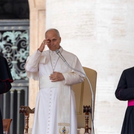 Pope in white robe salutes at a podium, flanked by two black-and-magenta clergy in a solemn ceremony.