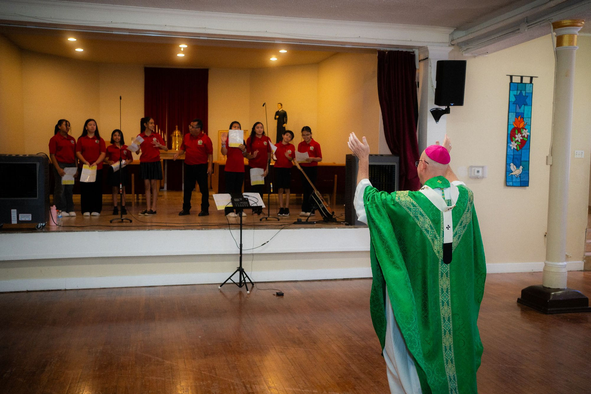 Bishop in green vestments blesses a choir in red shirts on a church stage, with microphones and warm sanctuary lighting.