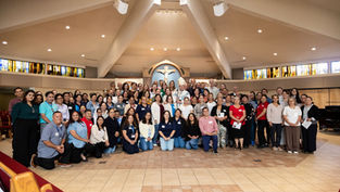 Large group poses in a church sanctuary with stained-glass windows, a bishop, and a crucifix centered behind them.