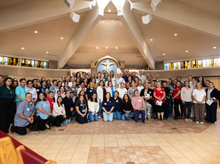 Large group poses in a church sanctuary with stained-glass windows, a bishop, and a crucifix centered behind them.