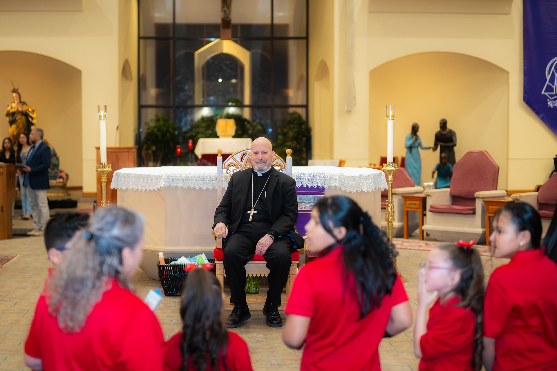 A man in black robes sits on a chair in a church, facing children in red shirts. Background includes altar, candles, and statues.