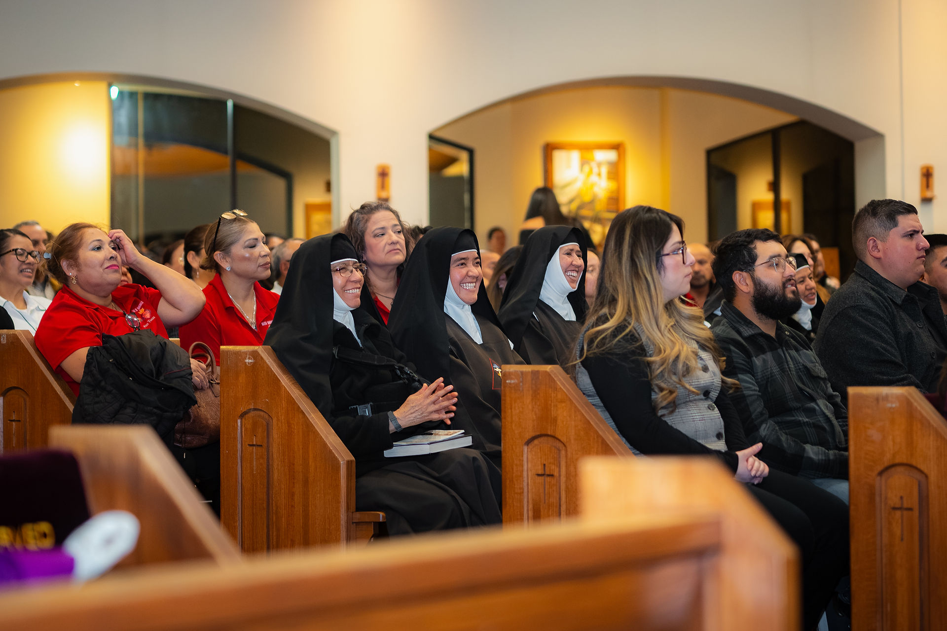 People seated in church pews, including nuns in habits and attendees in red shirts and dark clothing, smiling and listening attentively.
