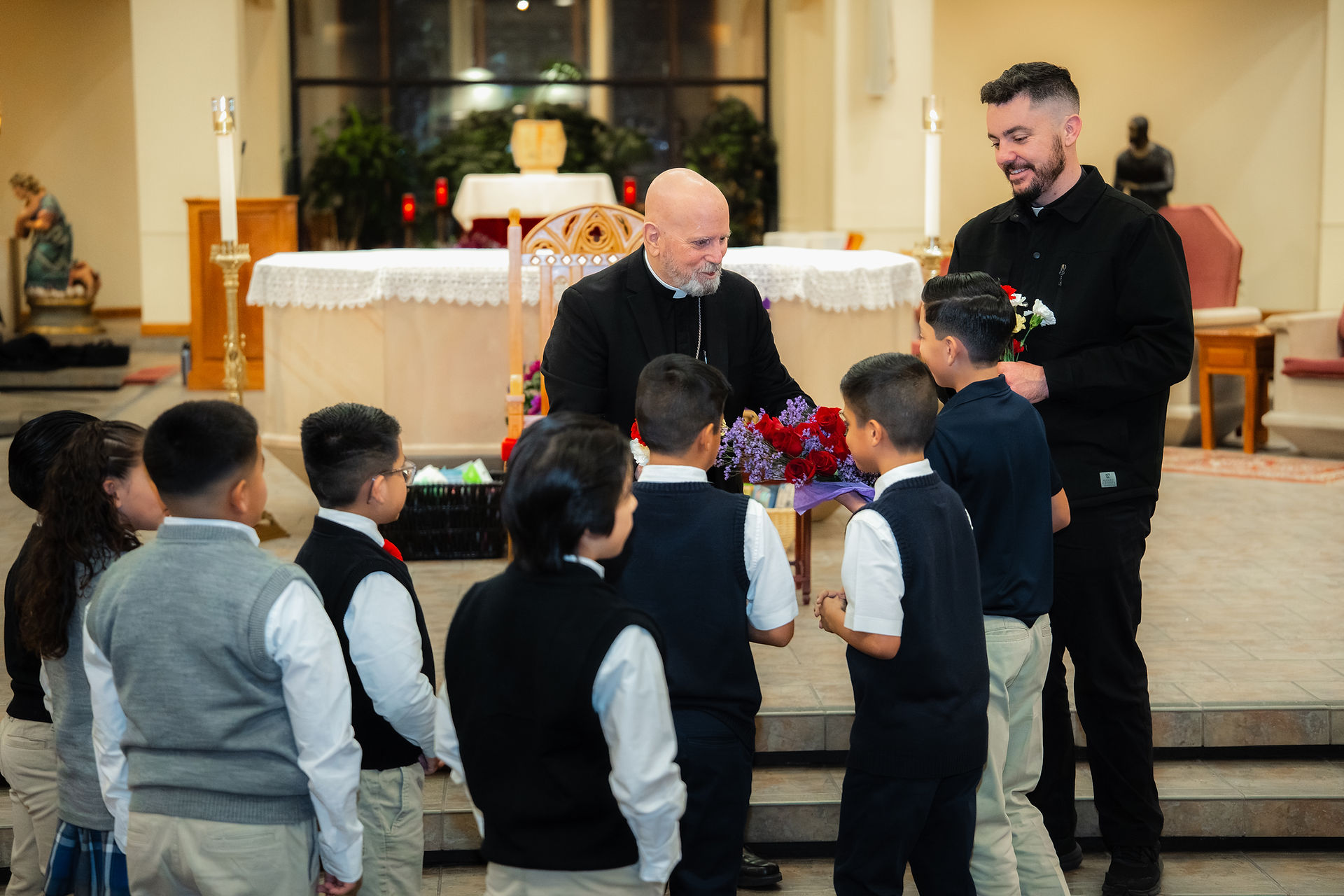Several Catholic school students present a bishop with flowers as a gift in a church. Another priest stands nearby, smiling. The mood is light and celebratory.