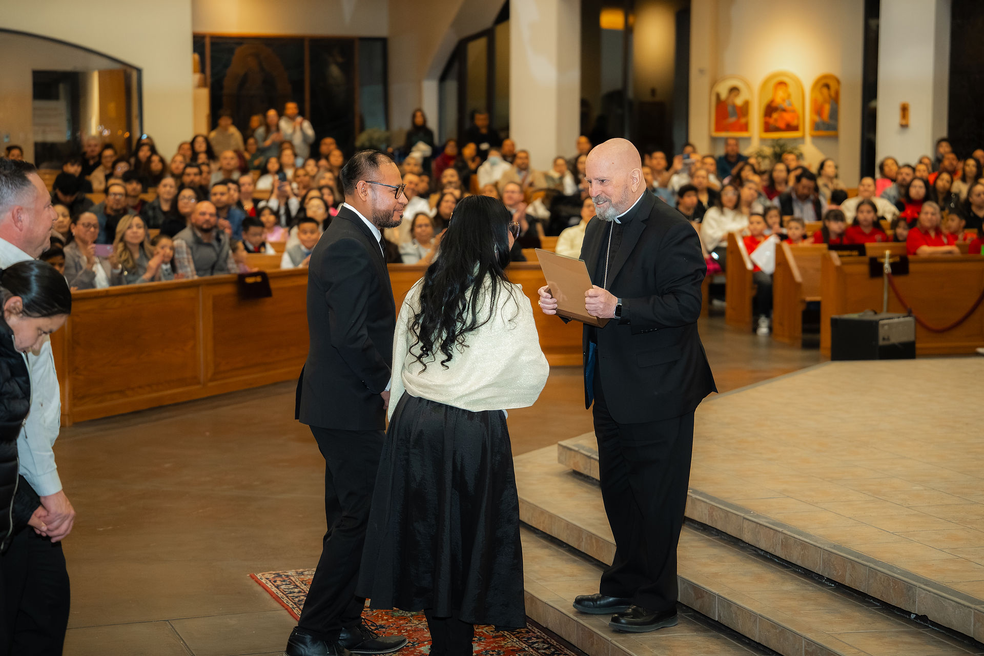 A bishop in black clerics receives a gift from adults in a church. The mood is light and celebratory.
