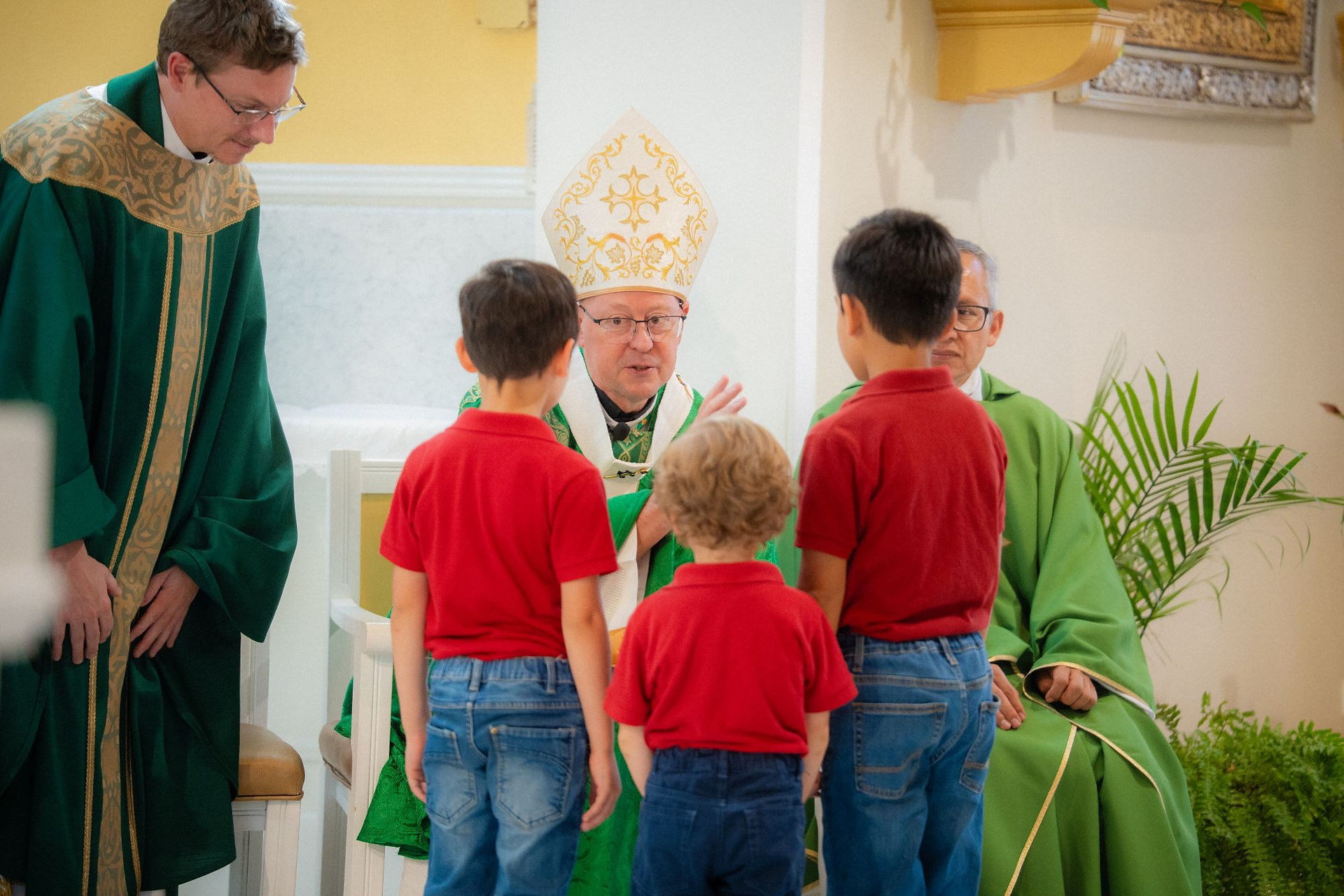 Bishop in white mitre speaks to three boys in red shirts during church service, with priests in green vestments nearby.
