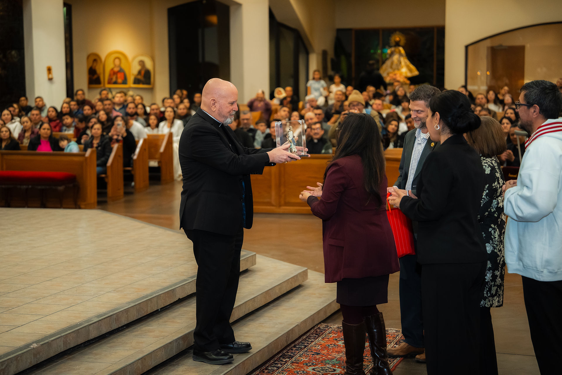 A bishop in a black suit receives a statue from several adults in a church. Congregation visible in the background. The mood is celebratory.