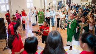 Church congregation claps and sings around a guitarist, with priests in green vestments in a bright hall.