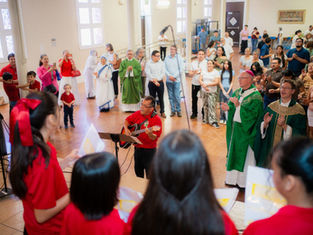 Church congregation claps and sings around a guitarist, with priests in green vestments in a bright hall.