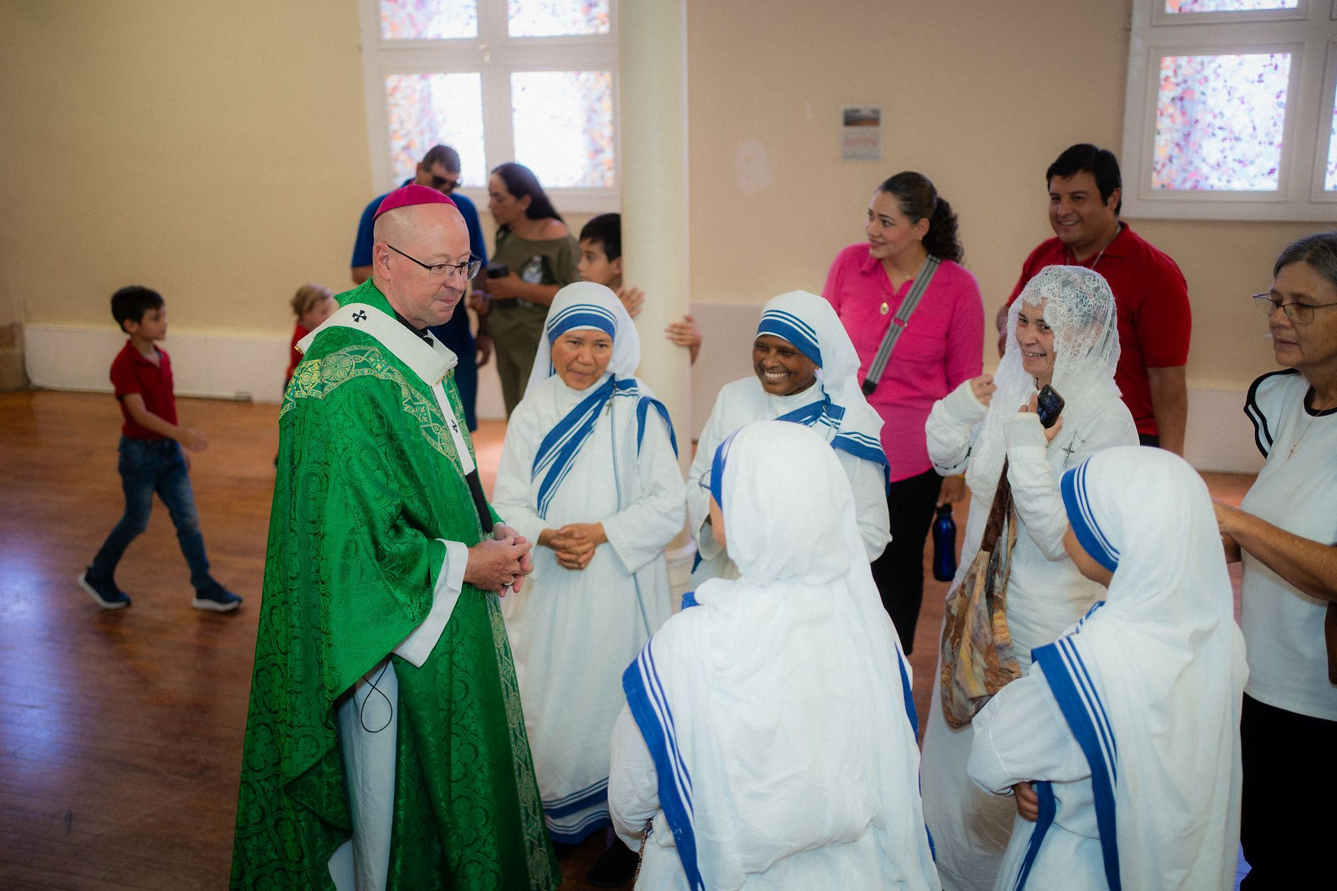 Catholic bishop in green vestments talks with smiling nuns in white-and-blue habits in a bright hall, with onlookers behind them.