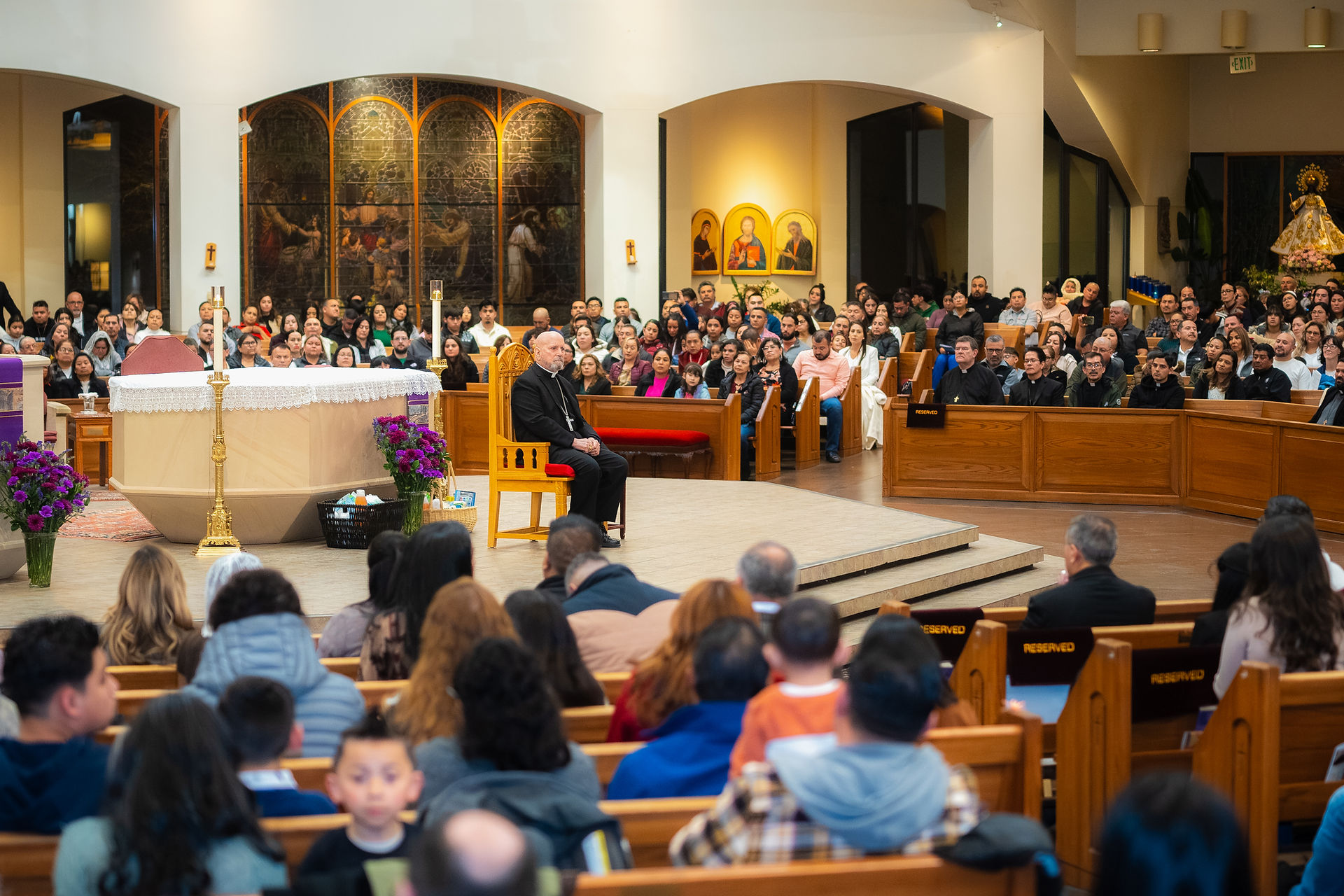 Clergy sits by altar in a crowded church. Attendees watch, surrounded by floral arrangements and stained-glass artwork. Reserved signs visible.