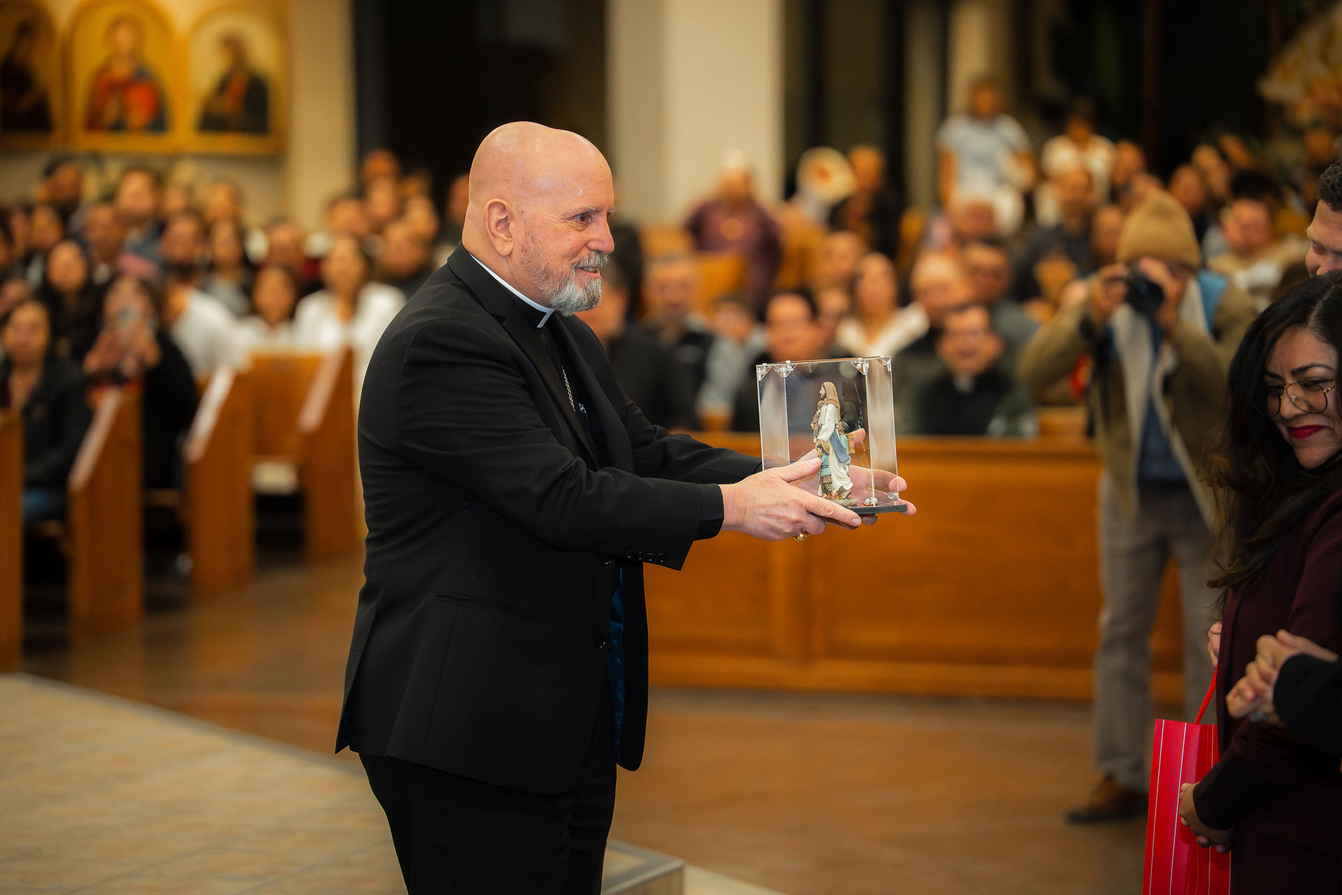 A bishop in a black suit receives a statue from several adults in a church. Congregation visible in the background. The mood is celebratory.