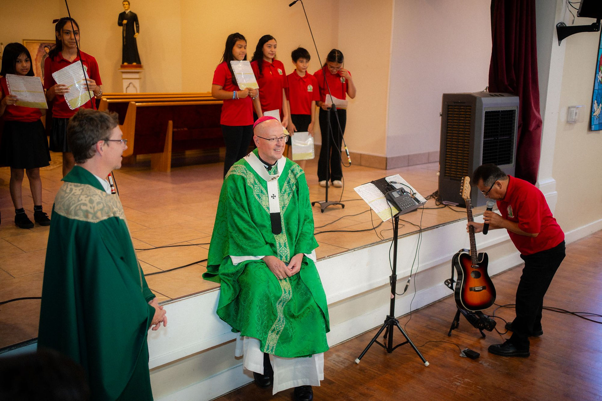 Bishop in green vestments smiles while children in red shirts hold sheets in a church; man tunes a guitar beside a microphone.
