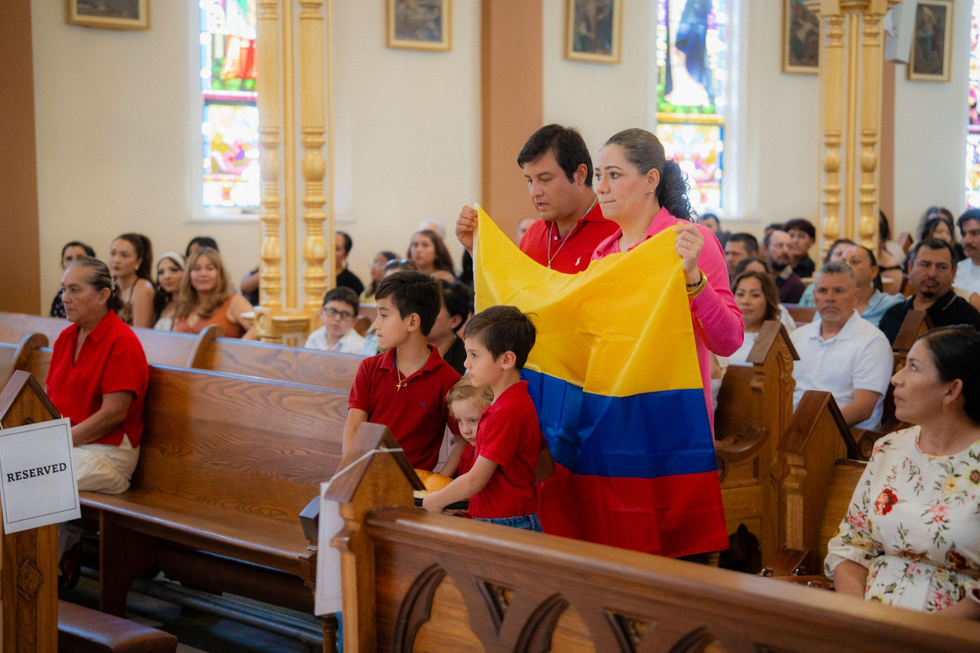 Family in red and pink stands in a church pew holding a Colombian flag, with a RESERVED sign and stained-glass windows behind them.