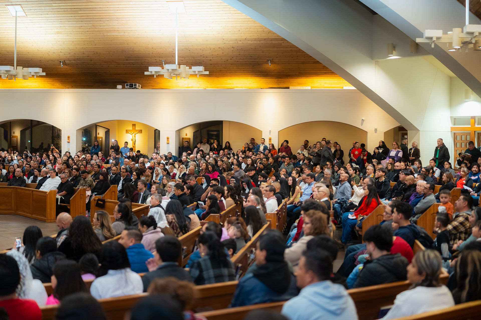 A full church during a religious service. A crucifix is visible in the background. The mood is warm and prayerful.