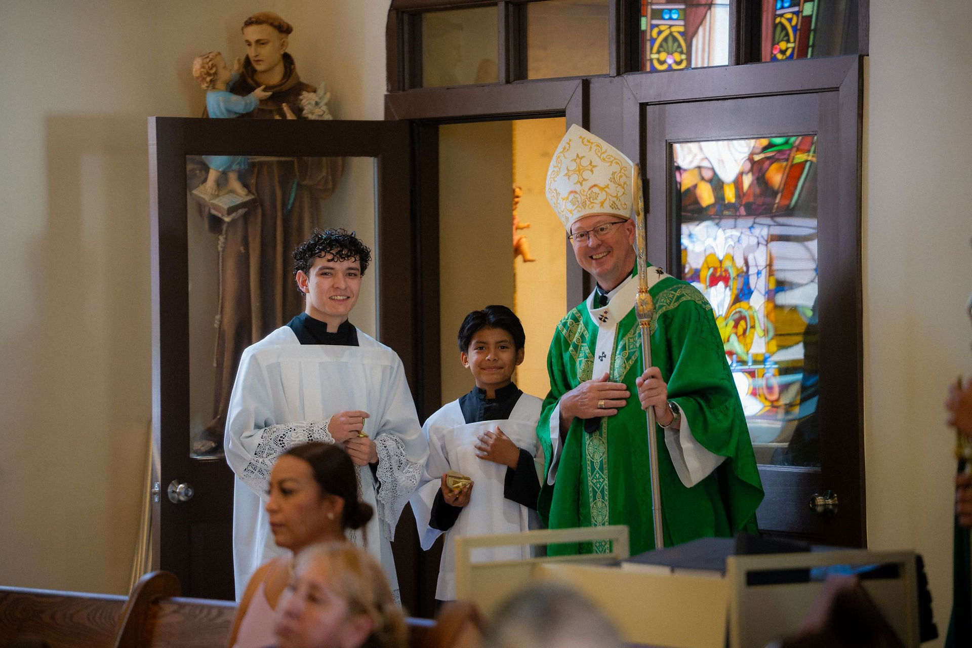 Smiling bishop in green vestments stands with two altar servers in a church doorway, with stained glass behind.