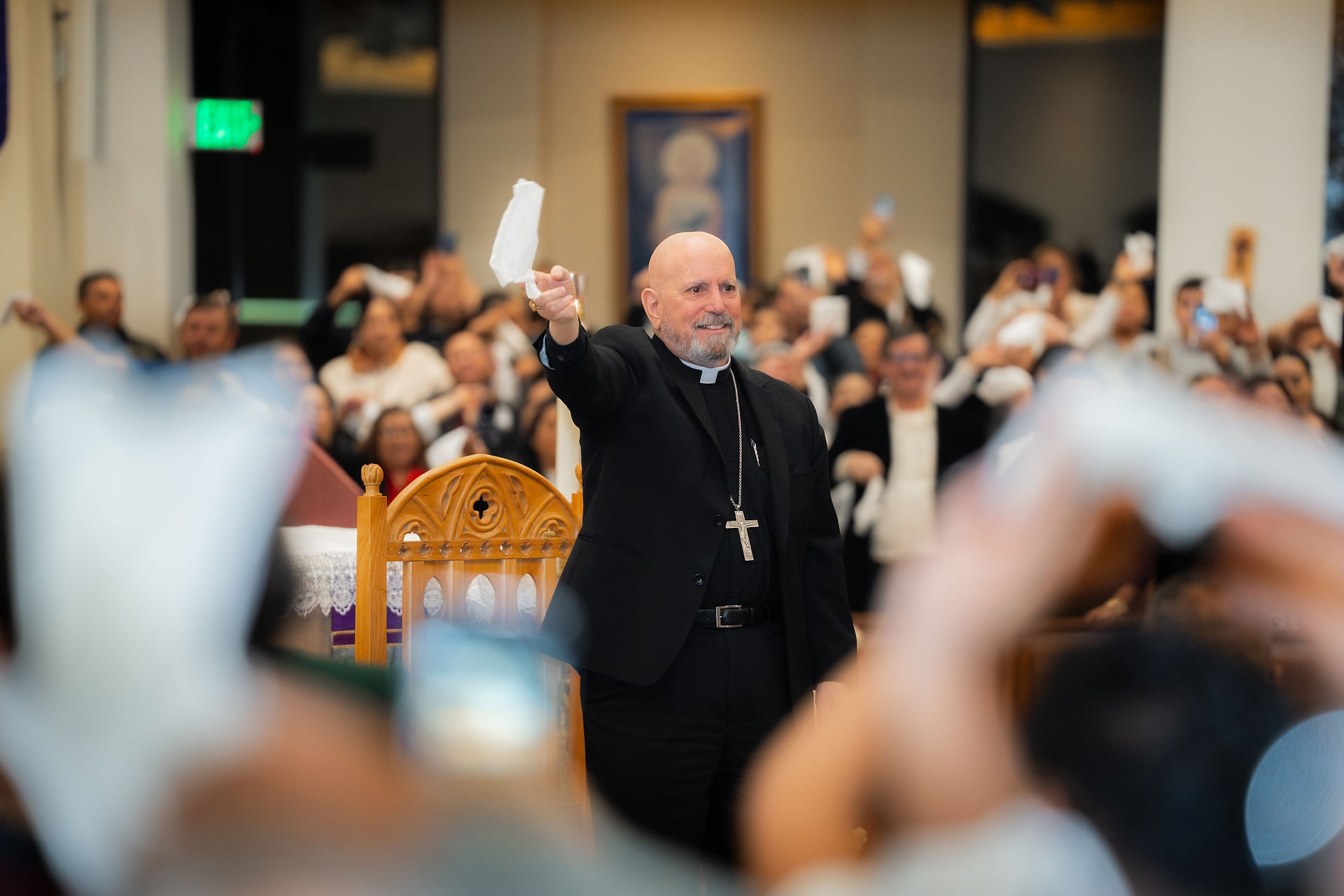 A clergyman in a black suit and cross necklace raises a handkerchief in a crowded church. People mimic the gesture, creating a festive mood.