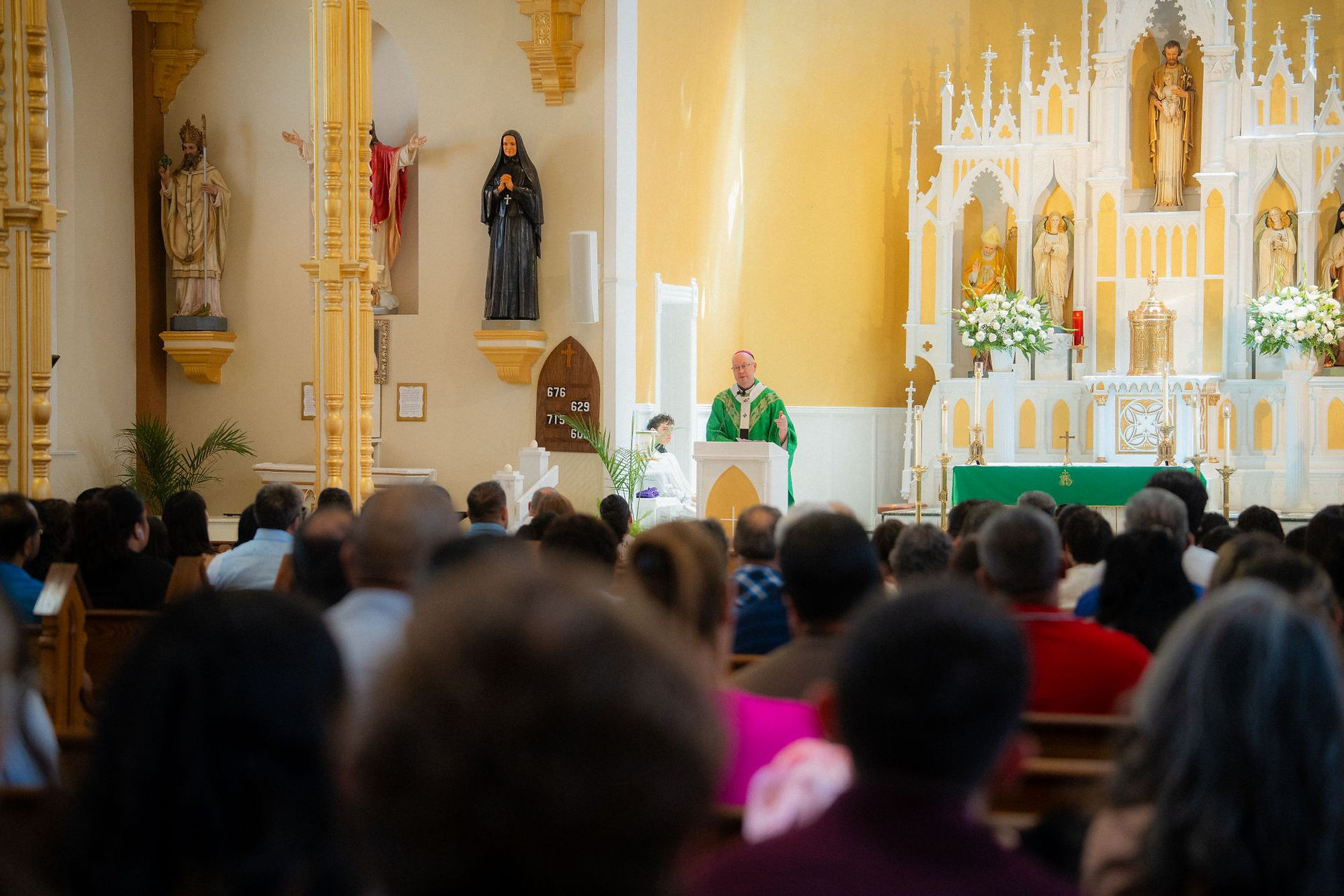 Bishop in green vestments speaks at church altar before seated congregation; hymn board shows 676, 629, 715.