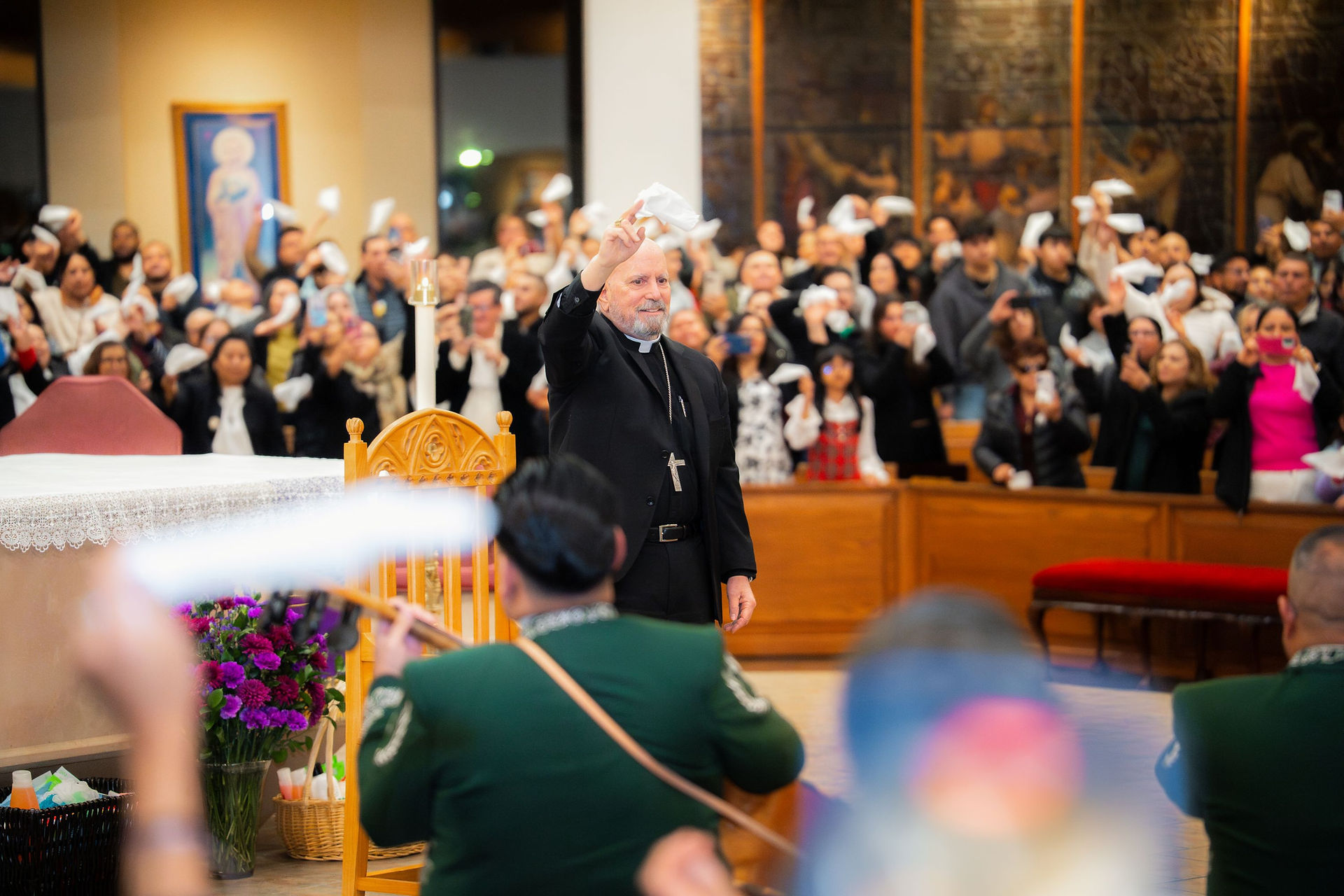 Bishop waving to a cheering congregation in a church. Crowd holds white cloths, vibrant flowers at altar. Joyful atmosphere.