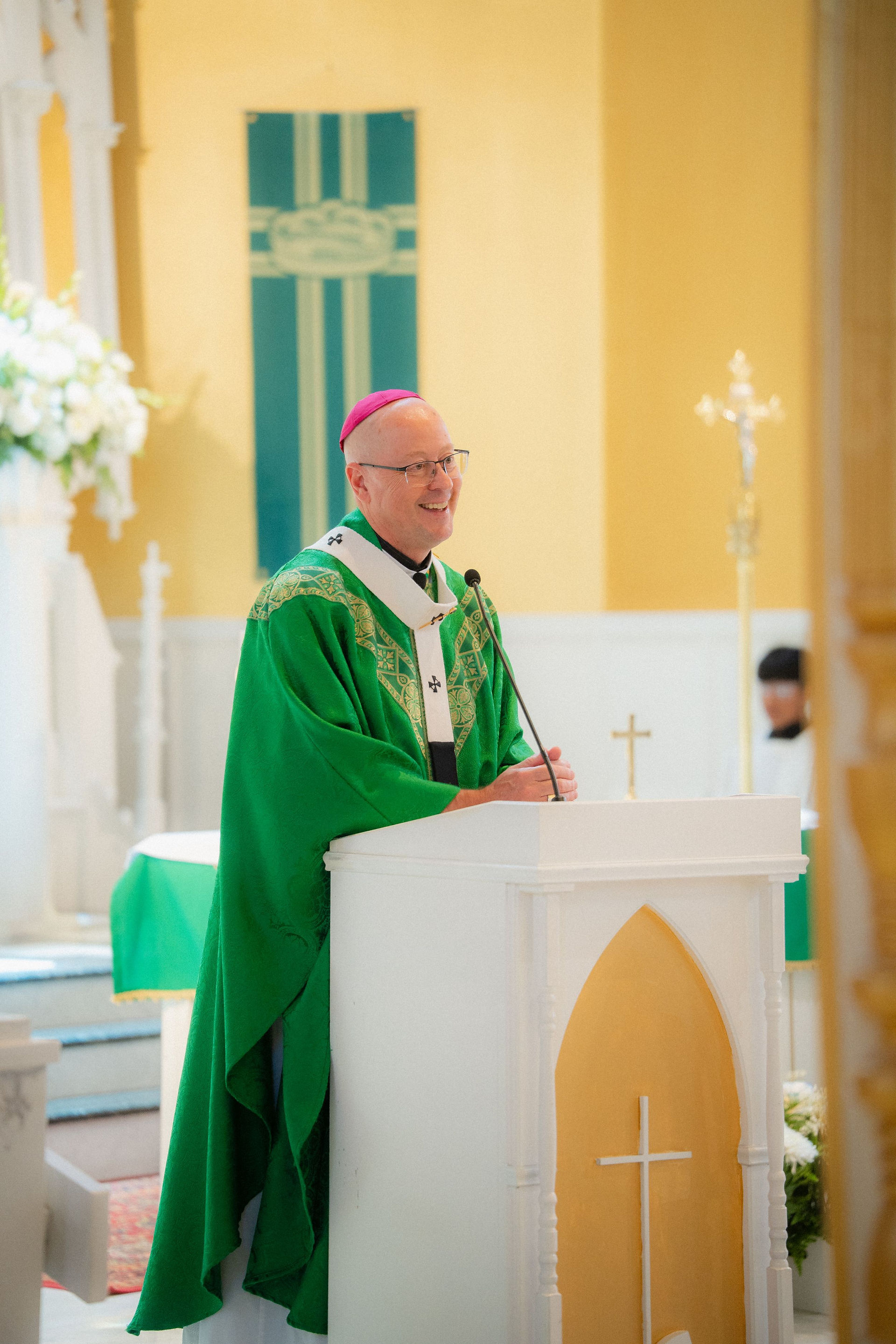Smiling bishop in green vestments speaks at a white church pulpit, with a cross and warm yellow sanctuary behind him.