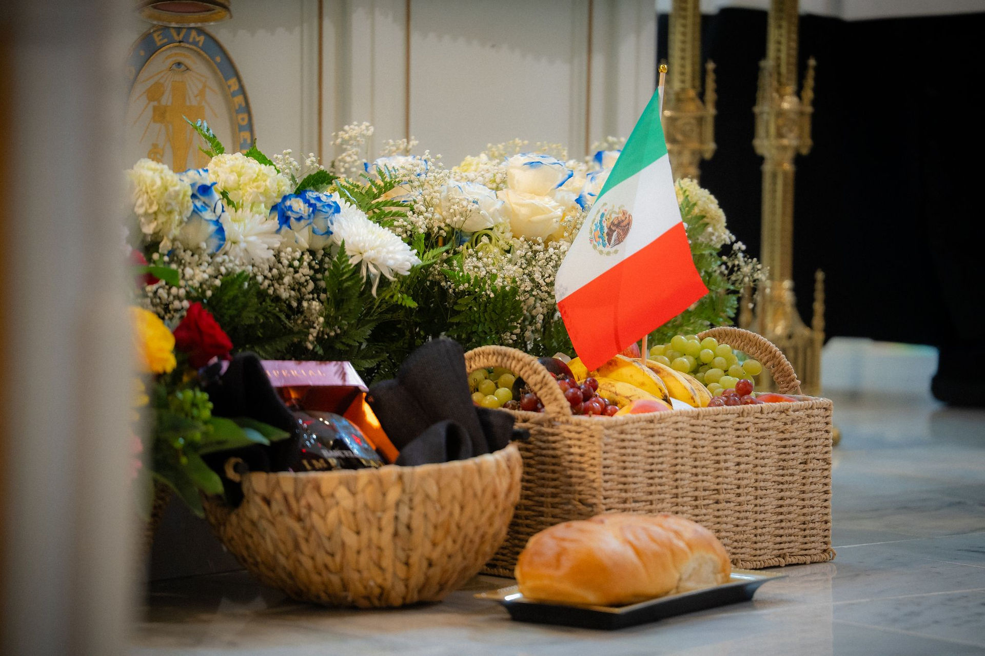 Fruit baskets, bread, and white-blue flowers with a small Mexican flag in a church, suggesting a solemn tribute.