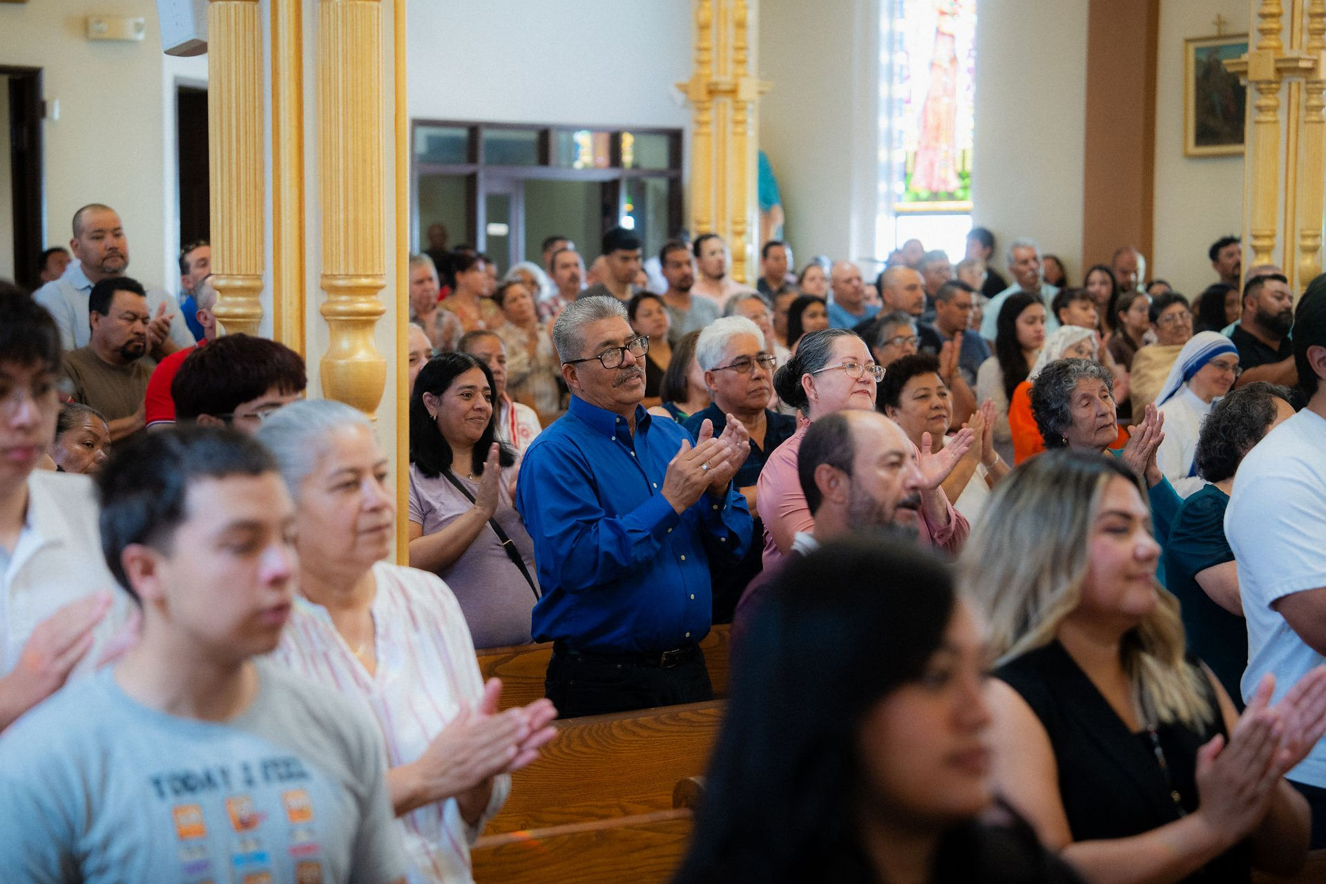Congregation applauds during a church service, standing in wooden pews beneath gold columns, with a warm, reverent mood.